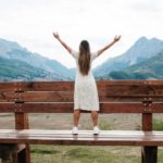 A woman stands on a huge oversized wooden park bench with her arms outstretched towards the mountainous horizon in the distance.