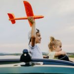 A smiling boy holds a toy plane aloft while a girl watches on from a car near an airport runway.
