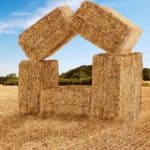 a young farmer stands back and admires his work in arranging bales of hay to form a house shape with two bales balancing against each other to form a roof, perched on bales tipped on their side in an abstract house shape on a freshly harvested paddock.