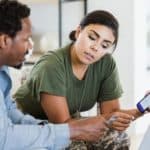 A couple sit in their home looking at a phone screen as if discussing a financial matter.