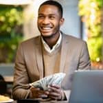 A man smiles as he holds bank notes in front of a laptop.