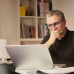 A senior investor wearing glasses sits at his desk and works on his ASX shares portfolio on his laptop.