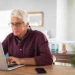 A senior investor wearing glasses sits at his desk and works on his ASX shares portfolio on his laptop2