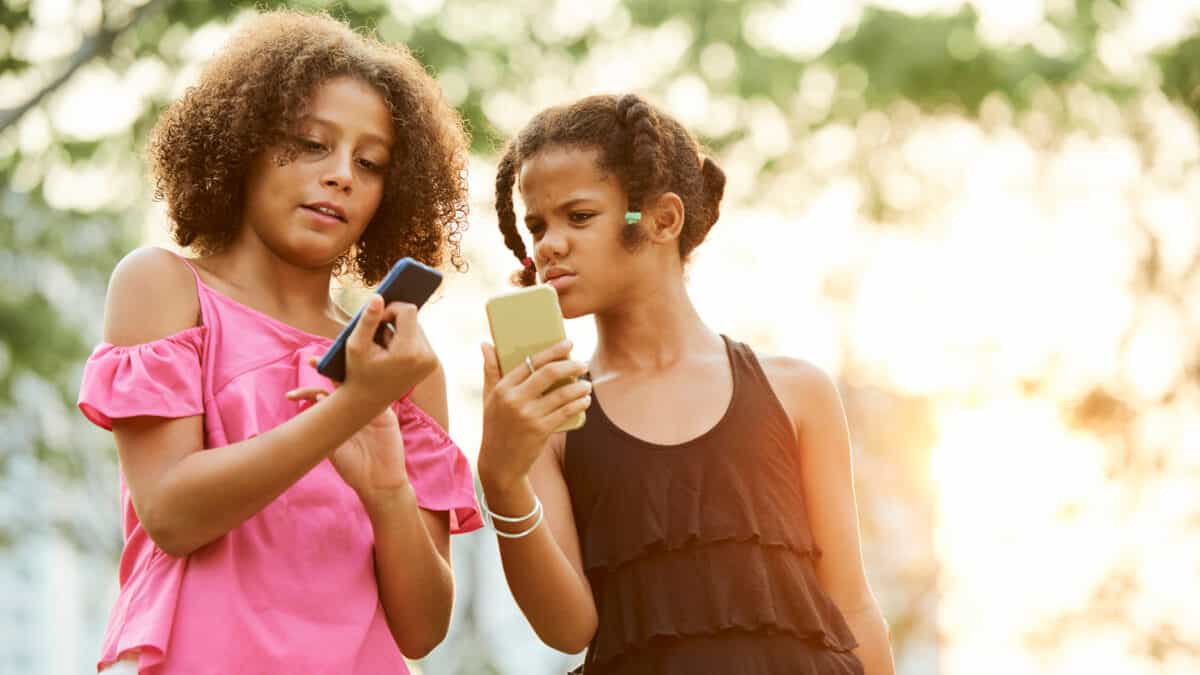 Confused African-American girls in casual clothing standing outdoors and comparing information on smartphones.