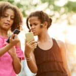 Confused African-American girls in casual clothing standing outdoors and comparing information on smartphones.