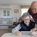 An older gentleman leans over his partner's shoulder as she looks at a tablet device while seated at a table in their classic Australian old person's home, complete with comfortable furniture and family photographs on the walls.