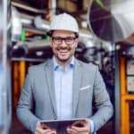 Smiling attractive caucasian supervisor in grey suit and with white helmet on head holding tablet while standing in power plant.