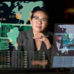 A woman sits at her desk thinking. She is surrounded by projections of world maps on various screens with data appearing below them.