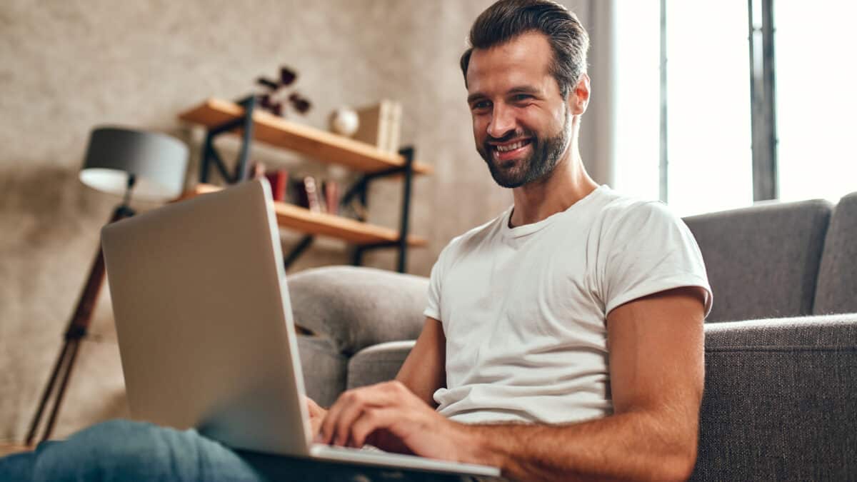 A man sits thoughtfully on the couch with a laptop on his lap.