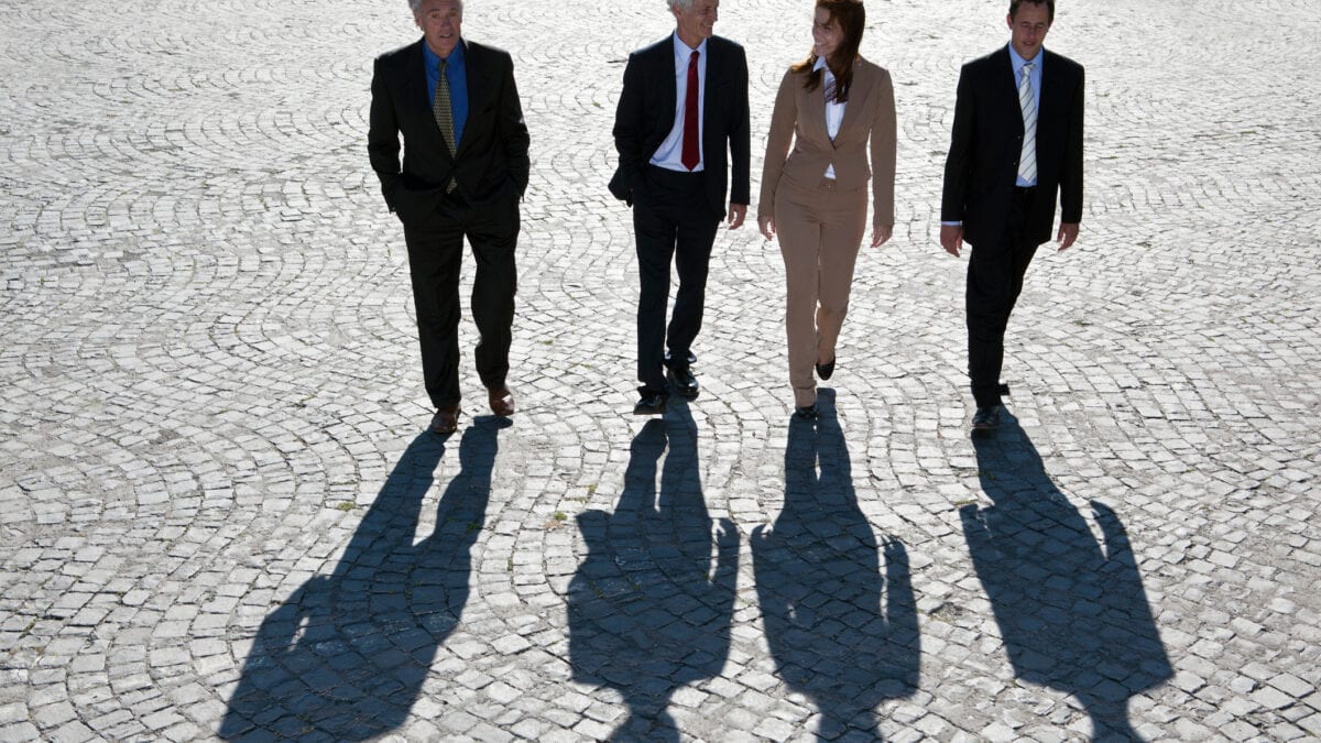 Four business people wearing formal business suits and ties walk abreast on a wide paved surface with their long shadows falling on the ground ahead of them.