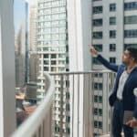 A man and a woman stand on an external balcony in a dense city environment filled with high rise buildings and commercial properties. The man is pointing up at a high rise building and the woman is looking on.