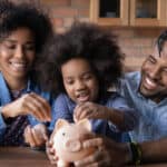 Young girl starting investing by putting a coin ion a piggybank while surrounded by her parents.