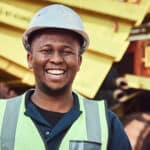 A young African mine worker is standing with a smile in front of a large haul dump truck wearing his personal protective wear.