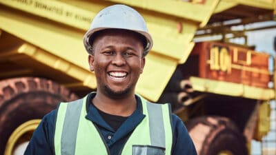 A young African mine worker is standing with a smile in front of a large haul dump truck wearing his personal protective wear.