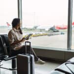 Woman on a tablet waiting in for her flight in an airport and looking through a window.