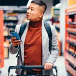 A man pushes a supermarket trolley with phone in hand down a supermarket aisle looking at the products on the shelves.