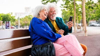 Married elderly man and woman in love spending time together on bench on a phone, symbolising retirement.
