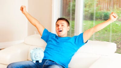 Man raising both his arms in the air with a piggy bank on his lap, symbolising a record high.
