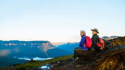 An older couple of retirement age and wearing hiking gear sit on a rocky outcrop gazing out at a sensational view of a rock formation and a waterway in the Australian bush.