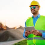 Image of young successful engineer, with blueprints, notepad and digital tablet, observing the project implementation on construction site and in mine.