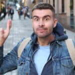 A young man wearing a backpack in a city street crosses his fingers and hopes for the best.