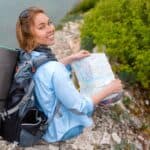 A smiling woman with backpack and a map sits on a rocky cliff about to embark on a new investing journey.