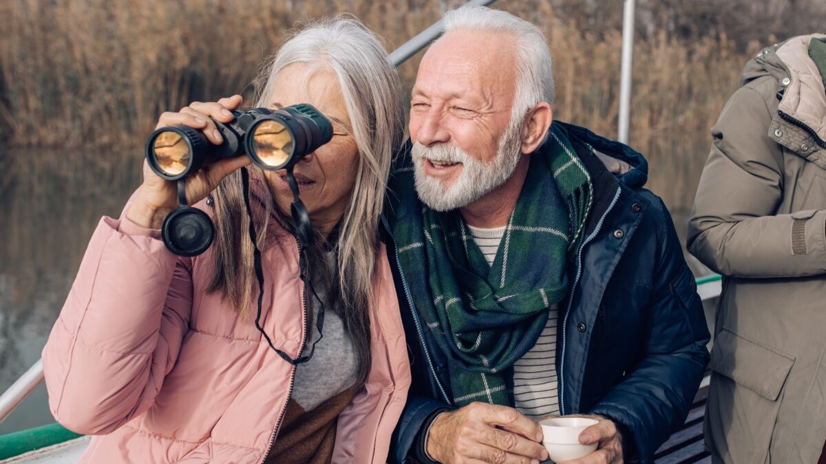 A happy elderly couple enjoy a cuppa outdoors as the woman looks through binoculars.