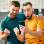 Two happy excited friends in euphoria mood after winning in a bet with a smartphone in hand.