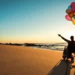 A joyful woman in a wheelchair on a beach holds a bunch of colourful balloons and spreads her arms wide towards the sunset.