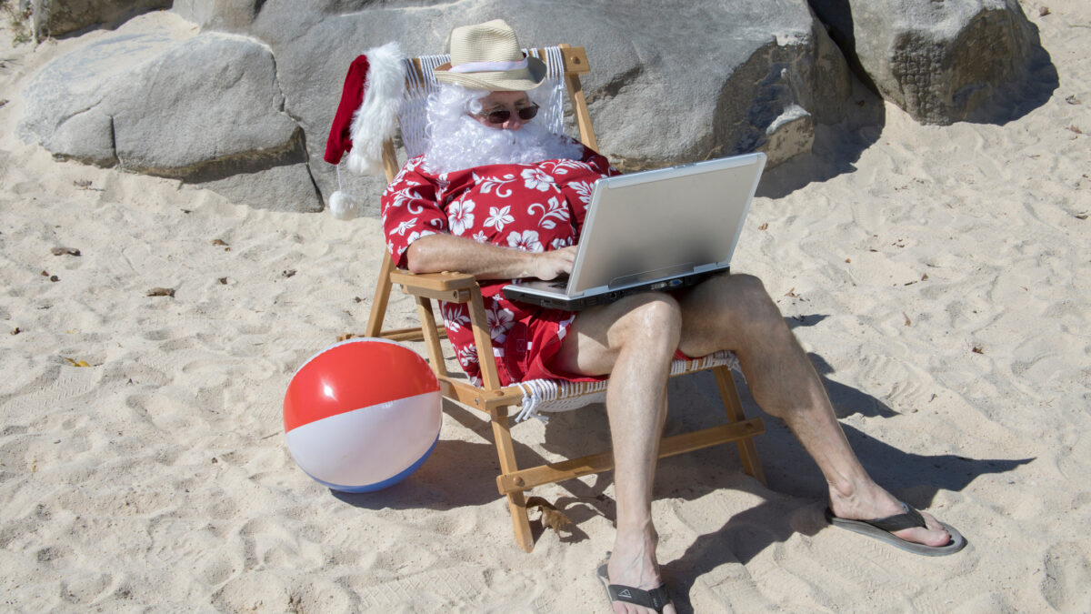 A fun depiction of summer Santa Claus -- wearing red swimming trunks and Hawaiian shirt -- sitting in a deck chair on his laptop at the beach.