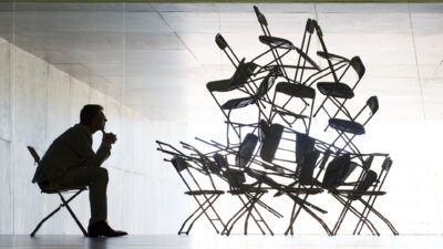 A businessman sits on a chair looking at a pile of chairs stacked up to the ceiling of a white empty room.
