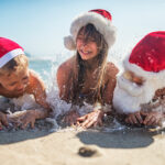 Three young people lie in the surf on a beach wearing santa hats.