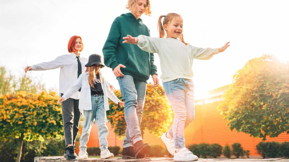 Smiling teenager boy and laughing girls show off their balancing skills by walking in a row on a wall in the autumnal sunny city park.