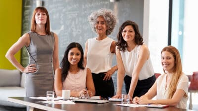 Five female colleagues at a work meeting, smiling to camera.