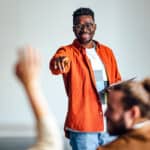 Cheerful man in a orange shirt standing in front of an audience holding a tablet and using hand gestures to interact with the audience.