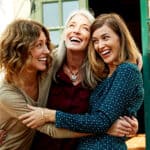 A mother and her two adult daughters embrace outdoors.