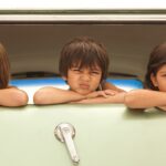 Three cute kids with mixed expressions poke their heads out from the back of a kombi.