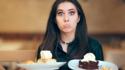 A bemused woman tries to choose between two slices of cake she holds on two plates.