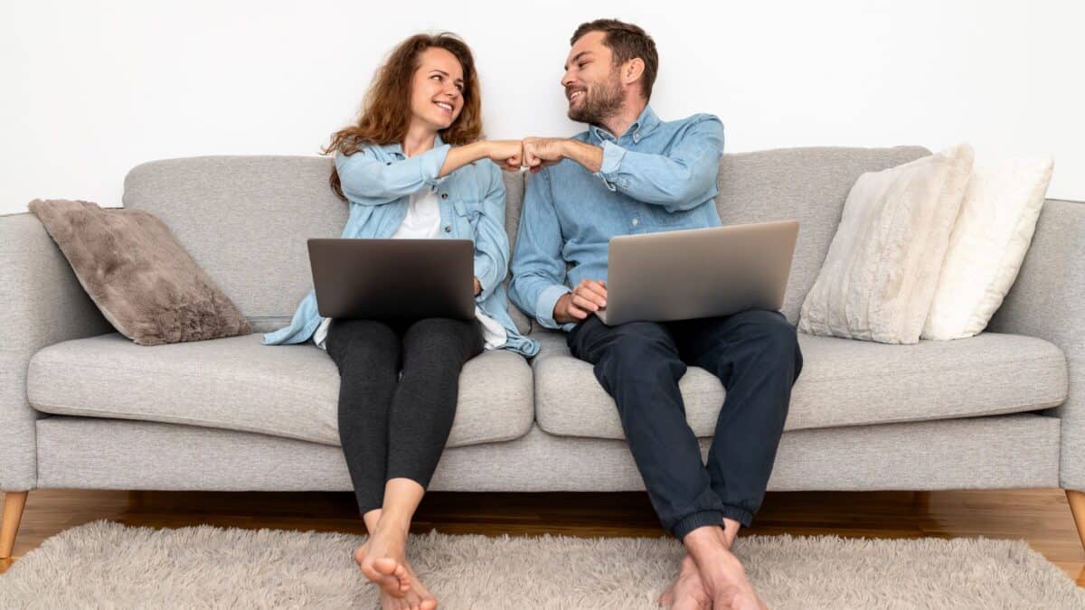 Smiling couple sitting on a couch with laptops fist pump each other.