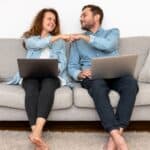 Smiling couple sitting on a couch with laptops fist pump each other.