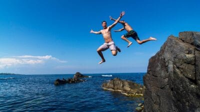 Two young risk-taking men pose for the camera as they jump off a cliff into the sea.