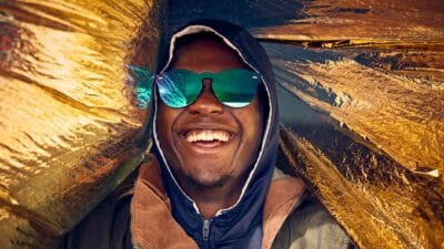 A cool man smiles as he is draped in gold cloth and wearing gold glasses.
