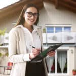 Happy woman standing in front of a house with a pen and clipboard.