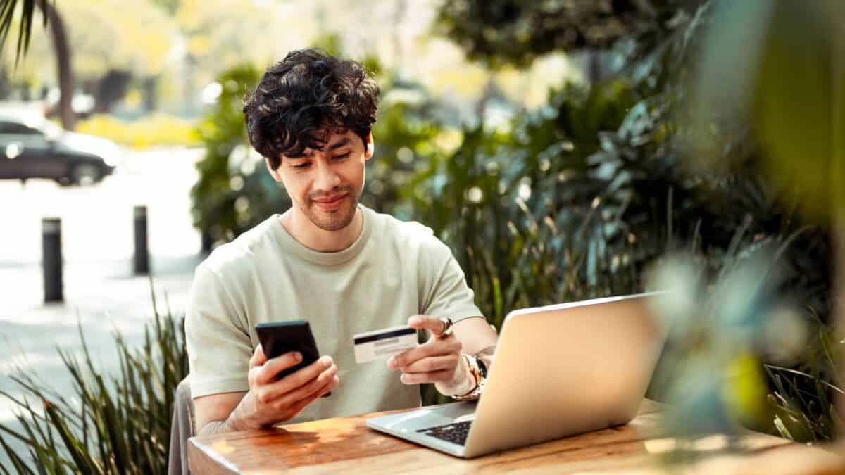 A young man sitting at an outside table uses a card to pay for his online shopping.