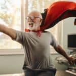 A happy elderly man wearing a red cape smiles as he jumps up like a hero from a massage table.