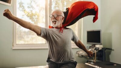 A happy elderly man wearing a red cape smiles as he jumps up like a hero from a massage table.