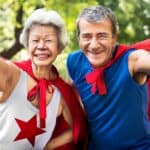 Two elderly people smiling with their fists pumping and with a cape on.