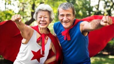 Two elderly people smiling with their fists pumping and with a cape on.