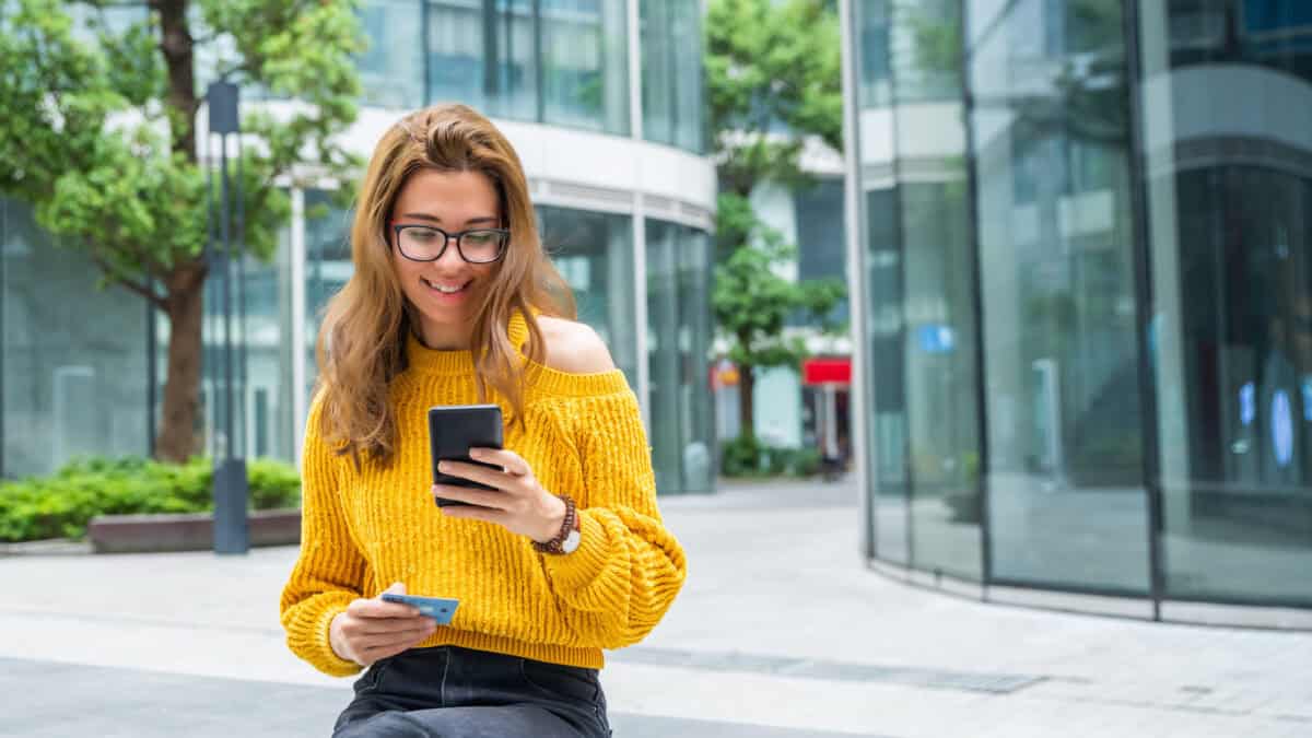 A young bank customer wearing a yellow jumper smiles as she checks her bank balance on her phone.