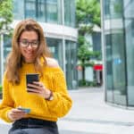 A young bank customer wearing a yellow jumper smiles as she checks her bank balance on her phone.